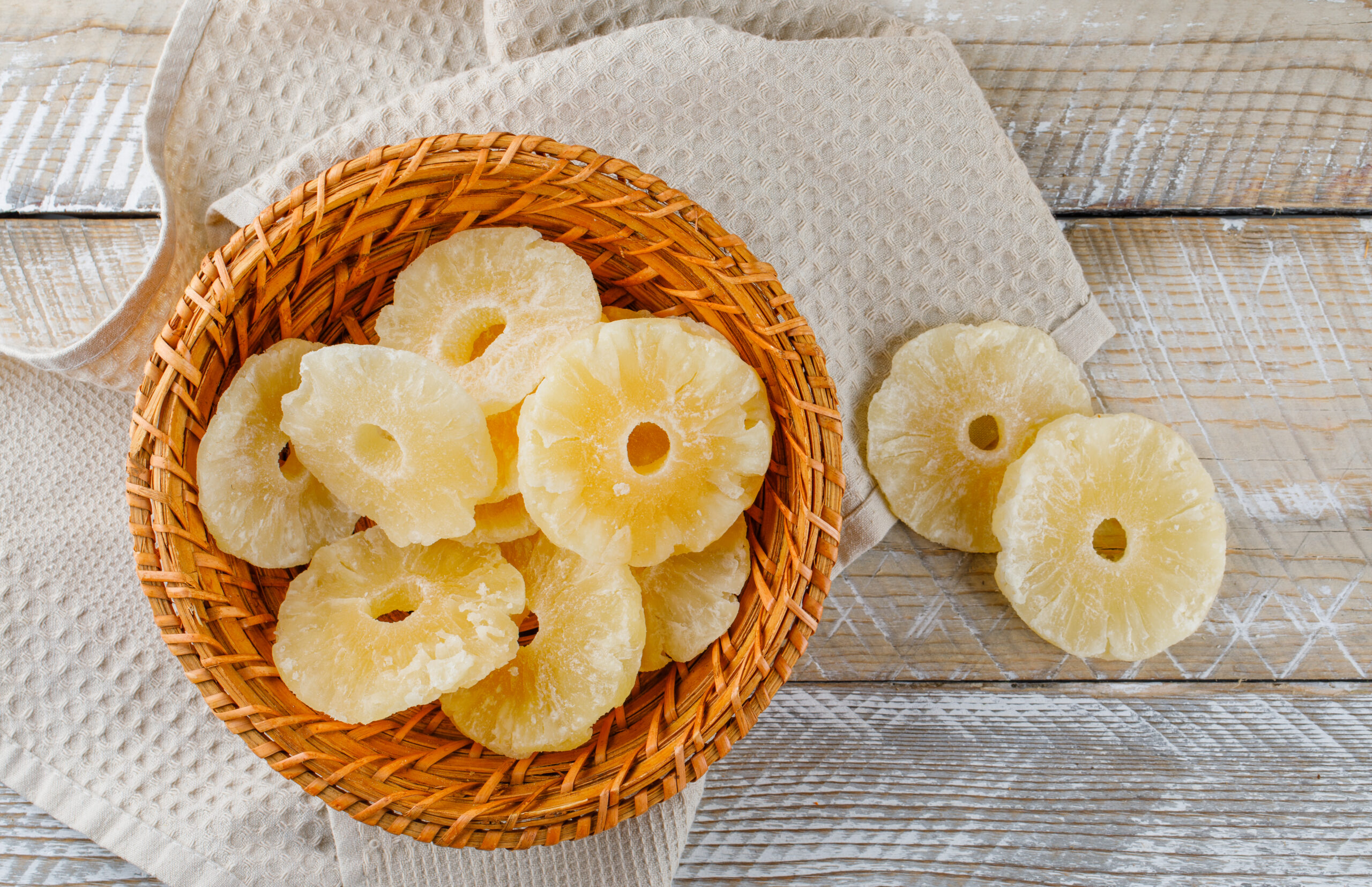 Dried pineapple rings in a wicker basket flat lay on wooden and kitchen towel background